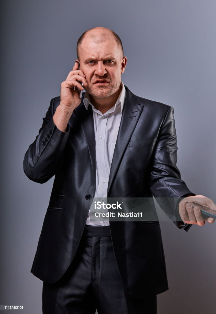 Angry not undestanding gloomy stressed angry business man talking on mobile phone very emotional in office suit on grey background. Closeup Angry not undestanding gloomy stressed angry business man talking on mobile phone very emotional in office suit on grey background. Closeup portrait Gangster Stock Photo
