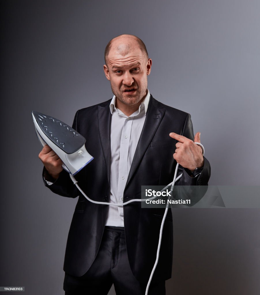 Bald fun gloomy comic business man holding the home iron and not want to ironing on grey background. Closeup Bald fun gloomy comic business man holding the home iron and not want to ironing on grey background. Closeup portrait Sulking Stock Photo