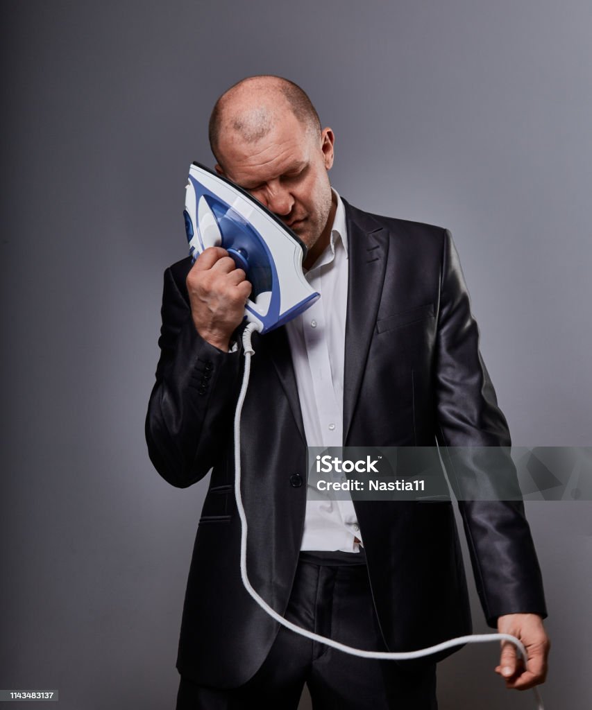 Bald fun tired unhappy loser failed business man punishing himself holding the home iron near the face in suit on grey background. Closeup Bald fun tired unhappy loser failed business man punishing himself holding the home iron near the face in suit on grey background. Closeup portrait Businessman Stock Photo