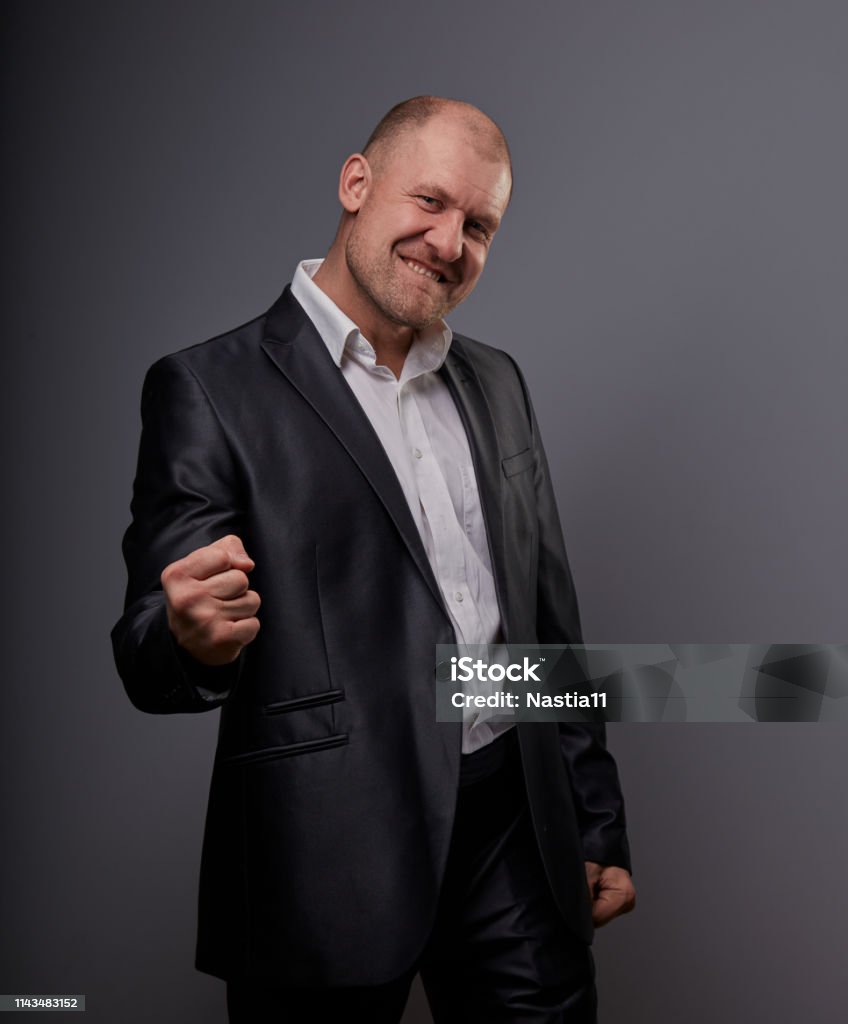 Exited bald business man in black suit showing the hands success winner sign on grey background. Closeup Exited bald business man in black suit showing the hands success winner sign on grey background. Closeup portrait Active Seniors Stock Photo