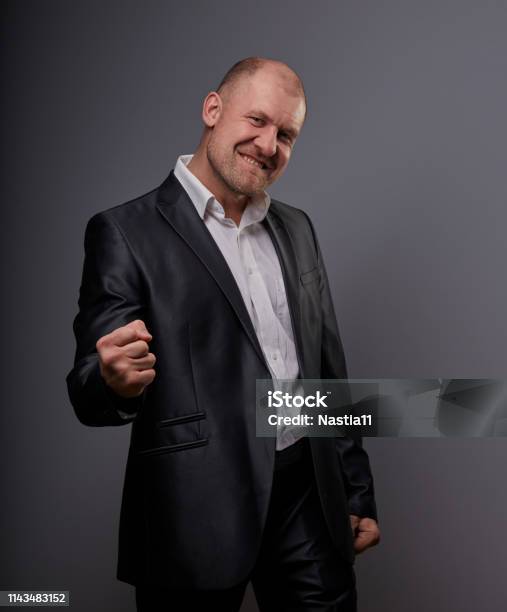 Exited Bald Business Man In Black Suit Showing The Hands Success Winner Sign On Grey Background Closeup Stock Photo - Download Image Now