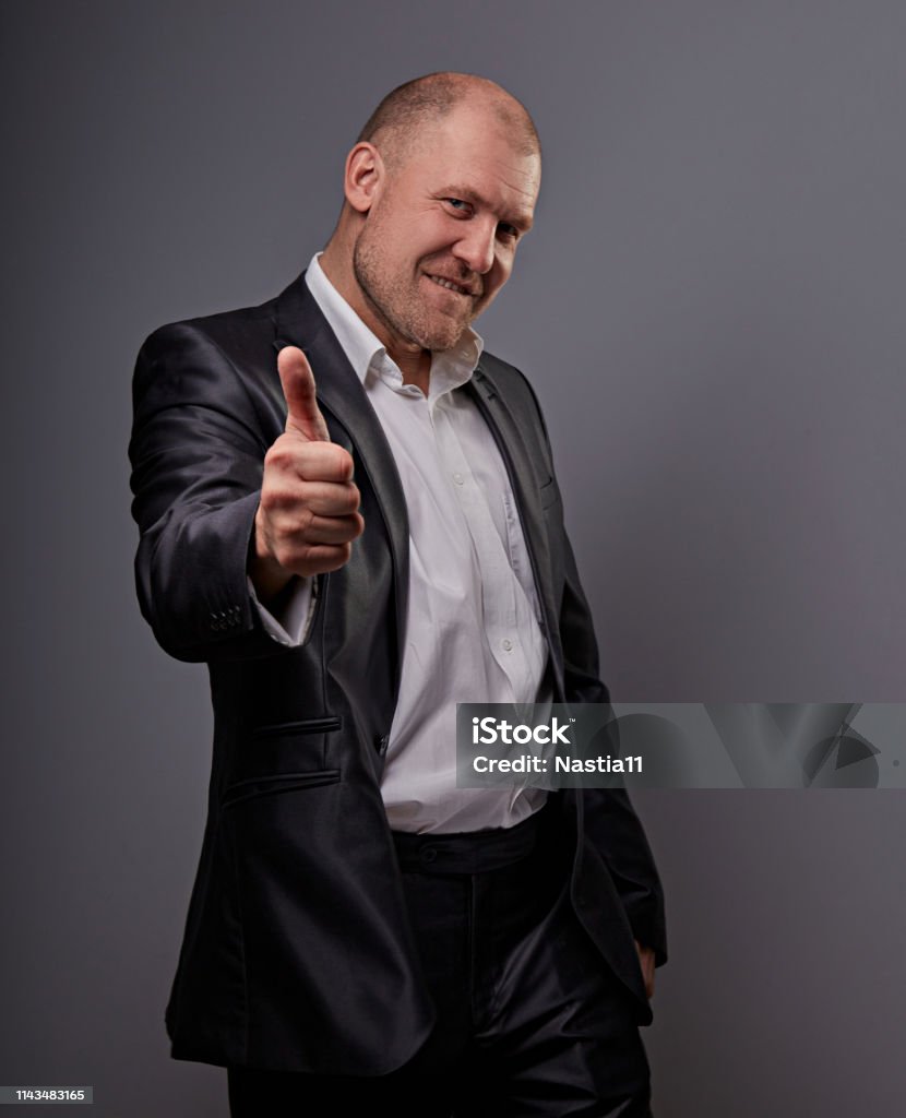 Exited comic bald business man in black suit showing the finger success thumb up sign on grey background. Closeup Exited comic bald business man in black suit showing the finger success thumb up sign on grey background. Closeup portrait Smiling Stock Photo