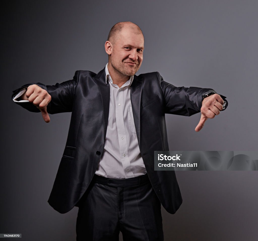 Fun comic bald business man in black suit showing the finger success thumb down sign on grey background. Closeup Fun comic bald business man in black suit showing the finger success thumb down sign on grey background. Closeup portrait Manager Stock Photo