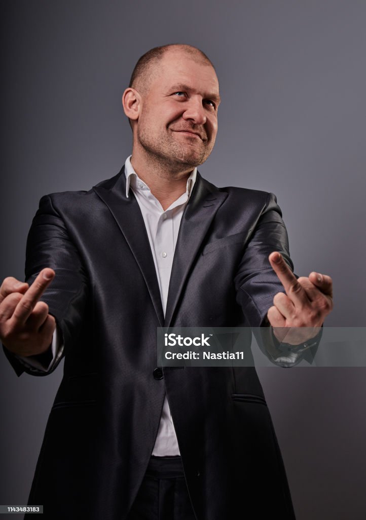 Fun comic bald business man in black suit showing the finger fuck sign on grey background. Closeup Fun comic bald business man in black suit showing the finger fuck sign on grey background. Closeup portrait Actor Stock Photo