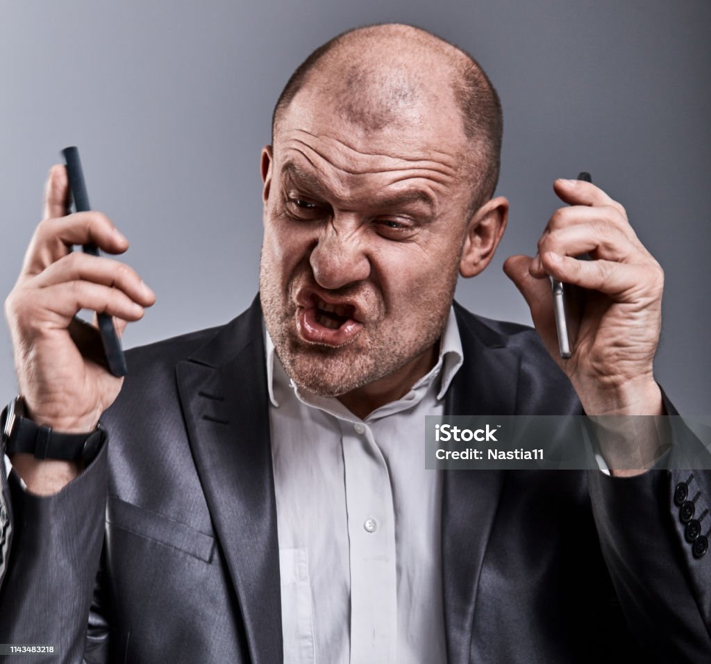 Turn nasty loud crying angry business man talking on two mobile phone very emotional in office suit on grey background. Closeup Turn nasty loud crying angry business man talking on two mobile phone very emotional in office suit on grey background. Closeup toned color portrait Adult Stock Photo