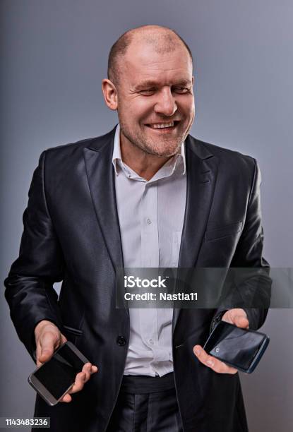 Positive Smiling Business Man Talking On Two Mobile Phones Very Emotional In Office Suit On Grey Studio Background Closeup Stock Photo - Download Image Now