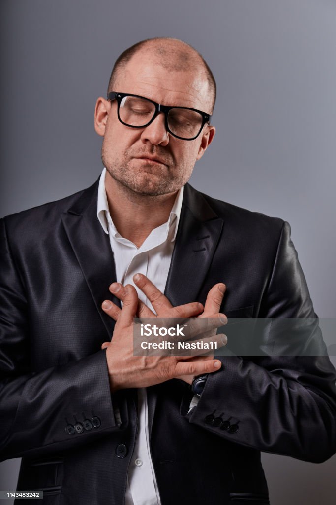 Thinking bald business man holding the chest two hands with serious face and closed eyes in eyeglasses in suit on grey background. Closeup Thinking bald business man holding the chest two hands with serious face and closed eyes in eyeglasses in suit on grey background. Closeup portrait 40-49 Years Stock Photo