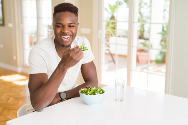 istock Handsome african young man eating a healthy vegetable salad using a fork to eat lettuce, happy and smiling sitting on the table 1143488582