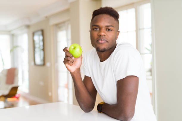 istock Young african american man eating fresh green apple with a confident expression on smart face thinking serious 1143488664