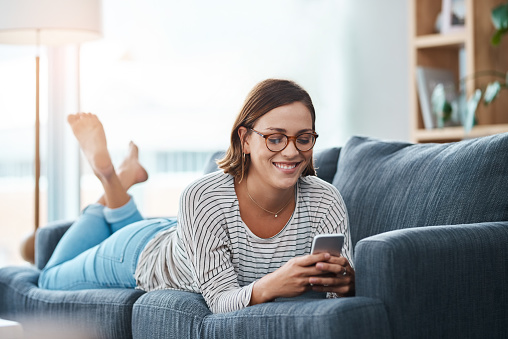 Full length shot of a happy young woman using her smartphone on the sofa at home