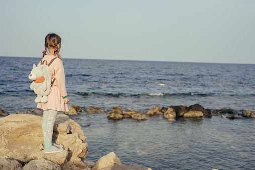 Little girl in is looking at the sea. She wears pink sweet-shot, jeans and blue backpack. Child stands very happy on the rocks. Concept of travelling, children holidays, treasure, tourism.