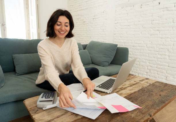 Happy housewife or entrepreneur woman paying bills, calculating costs, charges, mortgage, taxes with documents, laptop and calculator at home. In home and business finances and online banking concept. stock photo