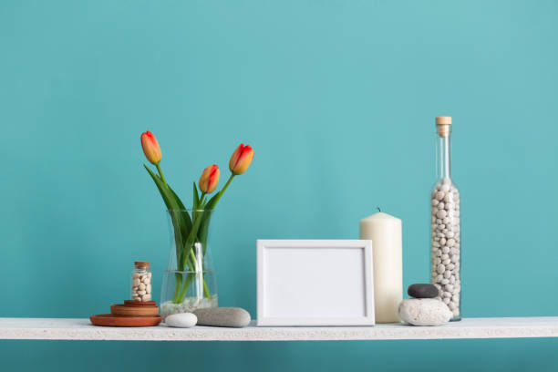 istock Modern room decoration with picture frame mockup. Shelf against turquoise wall with decorative candle, glass and rocks. Tulips in vase. 1143513183
