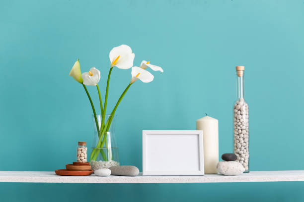 istock Modern room decoration with picture frame mockup. Shelf against turquoise wall with decorative candle, glass and rocks. Calla in vase. 1143513318