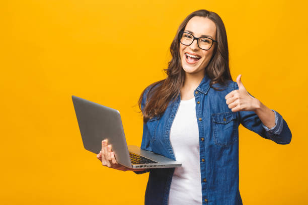 ラップトップコンピュータを保持し、黄色の背景上に孤立して親指を示す笑顔の若い美しい少女の肖像。 - holding laptop women computer ストックフォトと画像