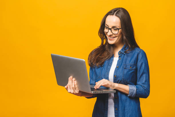 Portrait of a cheerful young woman wearing casual standing isolated over yellow background, holding laptop. Portrait of a cheerful young woman wearing casual standing isolated over yellow background, holding laptop. one person standing stock pictures, royalty-free photos & images