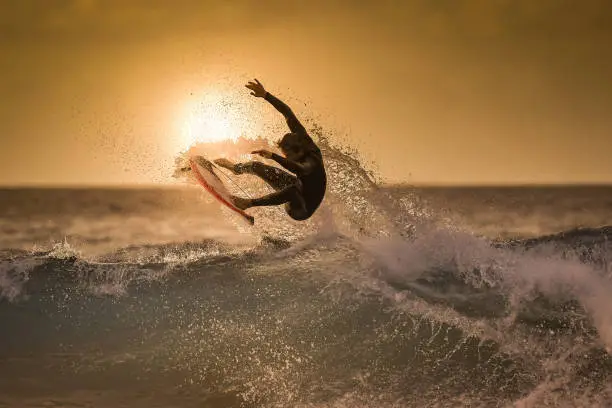 Photo of Surfer jumping on the wave at the golden hour on a tropical beach Sporty young man making tricks over the waves with a suf board Extreme sport and action in the water at the sunset with foam and spray