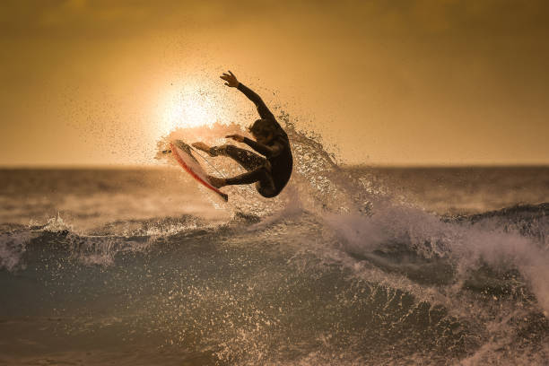 surfer jumping on the wave at the golden hour on a tropical beach sporty young man making tricks over the waves with a suf board extreme sport and action in the water at the sunset with foam and spray - surf imagens e fotografias de stock