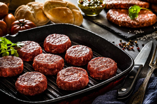 Several raw hamburger patties ready for cooking in a cast iron grill shot on dark kitchen table. Some ingredients for preparing and cooking hamburger like bread, tomatoes, pepper, salt garlic and some herbs are on the table. A slate board with raw hamburgers is at the top right. Predominant colors are red and black. Low key DSRL studio photo taken with Canon EOS 5D Mk II and Canon EF 100mm f/2.8L Macro IS USM.