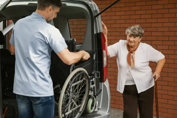 Caregiver holding wheelchair in the car for disabled elderly woman with walking stick