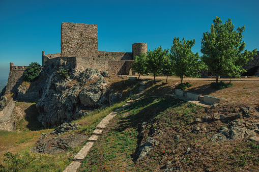 Stone walls and towers over rocky hill close to lawn garden with trees, in a sunny day at Castle of Marvao. An amazing medieval fortified village perched on a granite crag in eastern Portugal.