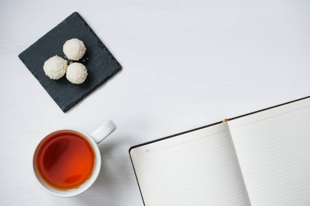 White coconut candy balls in plate shale dishes on rustic wooden background with a cup of tea and notebook.