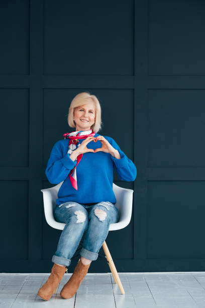 stylish senior woman heart shape hand care Stylish senior woman. Trendy blue outfit. Lady sitting on chair with heart shape hand sign. Care and happiness gesture. womens blue suede shoes stock pictures, royalty-free photos & images