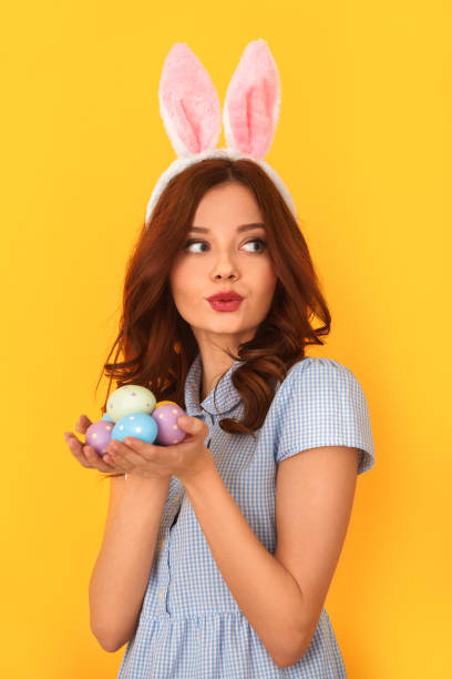 Young woman studio isolated on yellow wearing bunny ears holding eggs curious