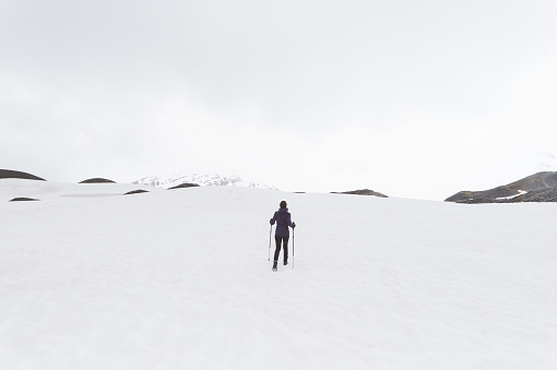 A young ethnic female hikes up an Alaskan glacier. It is snowy and foggy and there are mountains poking up over the ridge in front of her. She is using walking sticks. The shot is from behind.