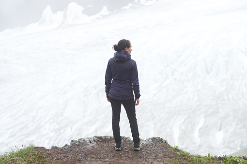 A young ethnic female hikes up an Alaskan glacier. It is snowy and foggy and there are mountains ahead of her as she stands at the edge of a cliff with her back to the camera and contemplates all the beauty.