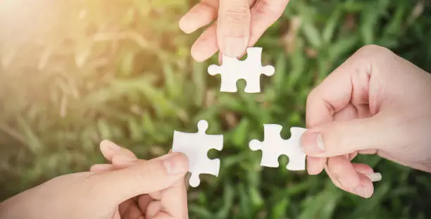 Hands trying to connect couple puzzle piece with sunset green grass background. Teamwork hands holding white jigsaw symbol of association and connection. Business strategy team together concept banner.