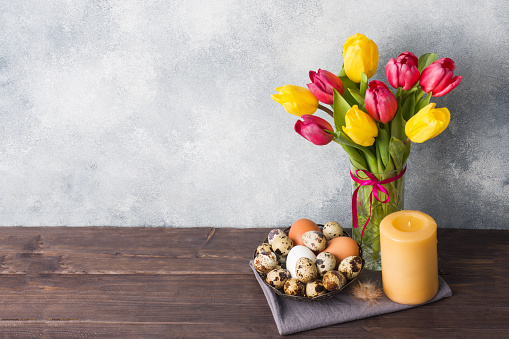 Concept Easter, tulips in a vase and a basket of eggs, a candle on a gray background. Copy space for product or text