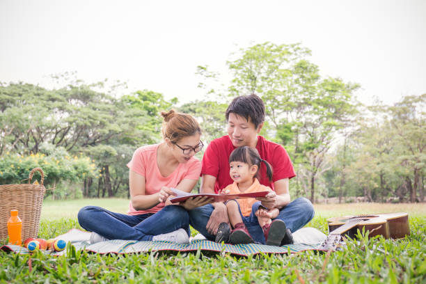 портрет счастливых азиатских родителей, читающих книгу с дочерью малыша. азиатский мужчина женщина читает историю маленькой девочке в пар� - baby spring child grass стоковые фото и изображения
