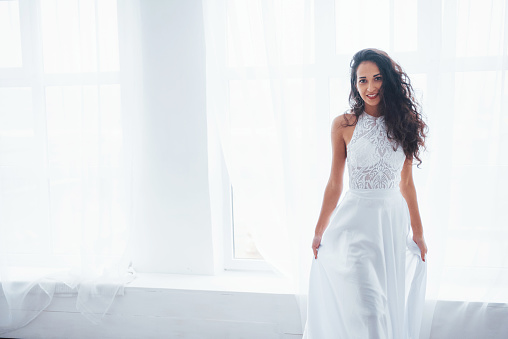 Luxury wear. Beautiful woman in white dress stands in white room with daylight through the windows.