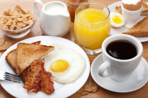 Stock photo showing close-up, elevated view of a white plate containing a fried breakfast with ramekin of baked beans, two sunny-side-up fried eggs, bacon rashers, sausages, grilled tomatoes, mushrooms and wholemeal toast.