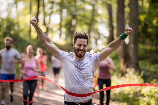 Cheerful athletic man running through finish line and celebrating victory with arms raised.