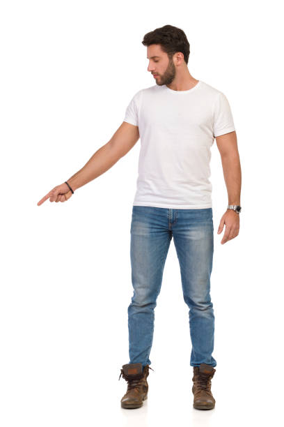 istock Serious Young Man In Jeans And White T-shirt Is Standing And Pointing Down. Front View 1143678056