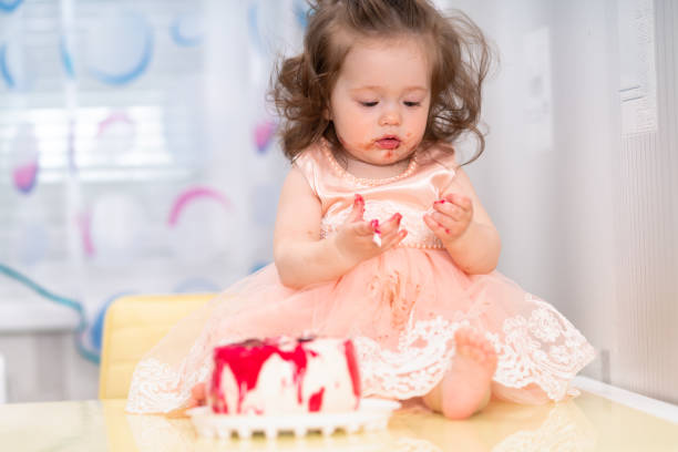 Cute little girl looking at her sticky hands as she sits eating a glazed cake for her birthday in a pink dress