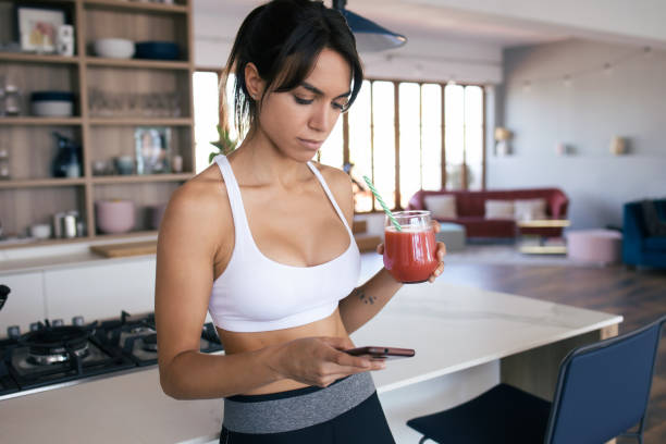 giovane donna sportiva che usa il suo cellulare mentre beve frullato di fragole in cucina a casa. - blueberry smoothie milk shake drink foto e immagini stock