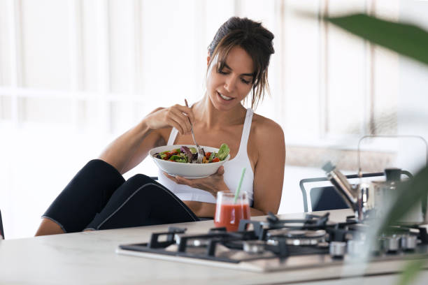 mujer joven deportiva comiendo ensalada y bebiendo jugo de fruta en la cocina en casa. - healthy eating sport exercising women fotografías e imágenes de stock