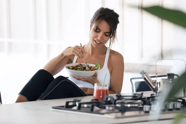 Photo of Sporty young woman eating salad and drinking fruit juice in the kitchen at home.