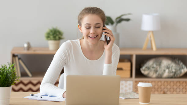 istock Smiling woman using laptop and talking on phone with friend, client 1143696562