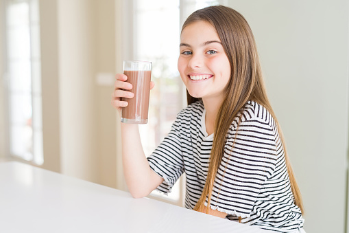 Beautiful young girl kid drinking fresh tasty chocolate milkshake as snack with a happy face standing and smiling with a confident smile showing teeth