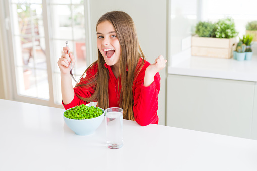 Beautiful young girl eating healthy green peas screaming proud and celebrating victory and success very excited, cheering emotion