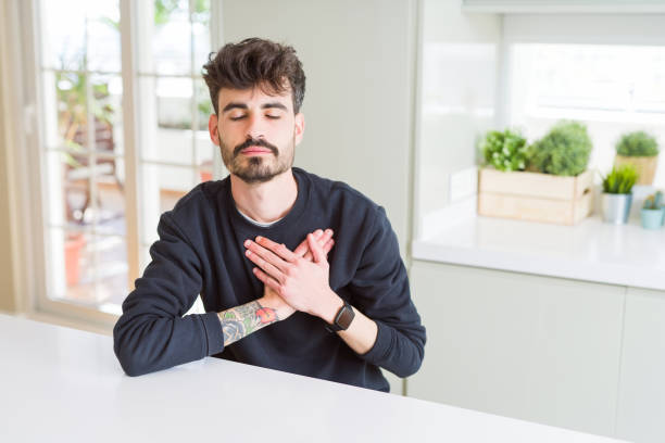 istock Young man wearing casual sweatshirt sitting on white table smiling with hands on chest with closed eyes and grateful gesture on face. Health concept. 1143706881