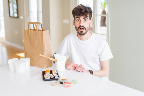 istock Young man eating asian sushi from home delivery afraid and shocked with surprise expression, fear and excited face. 1143707149