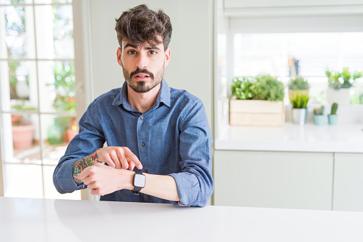Young man wearing casual shirt sitting on white table In hurry pointing to watch time, impatience, upset and angry for deadline delay