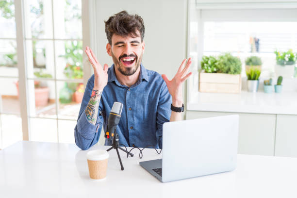 istock Young man recording podcast using microphone and laptop celebrating mad and crazy for success with arms raised and closed eyes screaming excited. Winner concept 1143707459