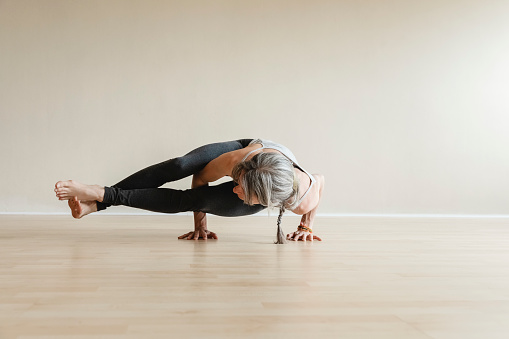 Adult woman wearing gray sportswear working out in indoor. Fit yogi woman doing Eight-angle pose, Astavakrasana.