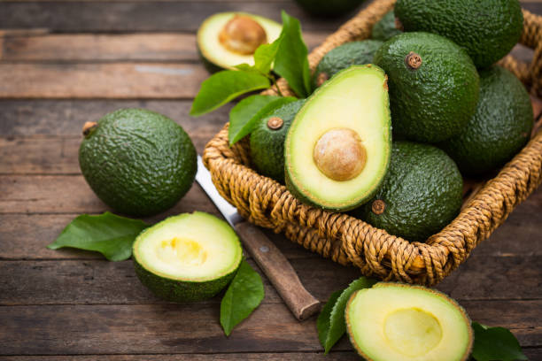 Close-up of a woman holding a ripe avocado while shopping at a local grocery market. Fresh green avocados are displayed in a basket, representing healthy eating, organic produce, and daily lifestyle choices focused on nutrition and sustainability.